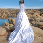 Novia con vestido blanco largo de espaldas levantando el ramo frente a rocas y un lago en Arizona