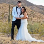 Pareja joven con traje blanco y vestido de novia posando en desierto de Arizona con cactus al fondo.