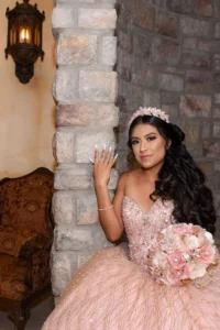 Quinceañera con vestido rosa y corona posando junto a columna de piedra, sosteniendo un ramo de flores decorado.