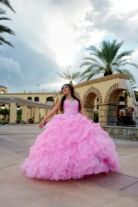 Quinceañera con vestido rosa esponjoso posando en exterior junto a una fuente y palmeras bajo cielo nublado.